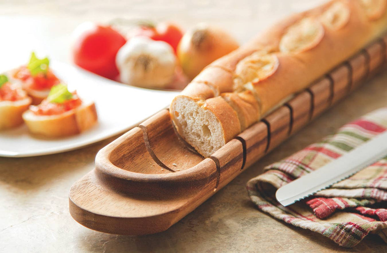 Wooden bread slicer with a loaf of bread on a table with a plate of appetizers.