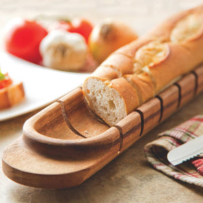 Wooden bread slicer with a loaf of bread on a table with a plate of appetizers.