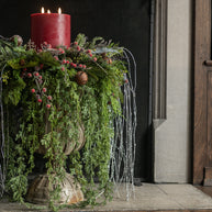 Decorative Christmas arrangement with red candle, greenery, and berries on a stone pedestal.