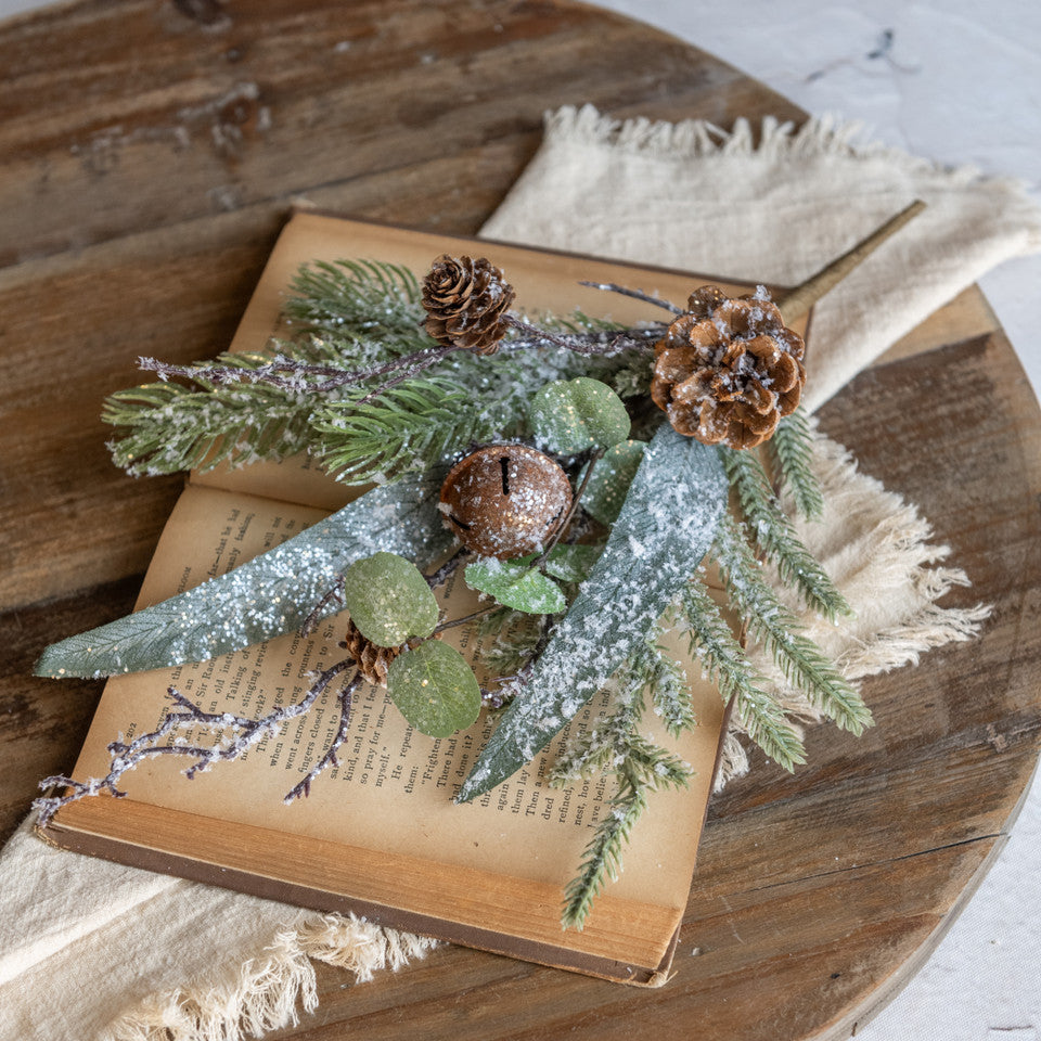 Decorative arrangement of greenery and pinecones on an open book with a wooden surface and beige fabric underneath.