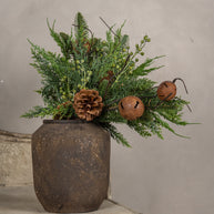 Decorative arrangement with greenery, pine cones, and bells in a rustic pot against a neutral background