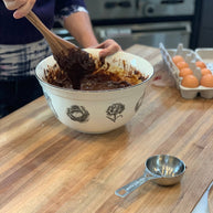 Person mixing chocolate batter in a bowl on a wooden countertop with eggs and measuring cup in the background.