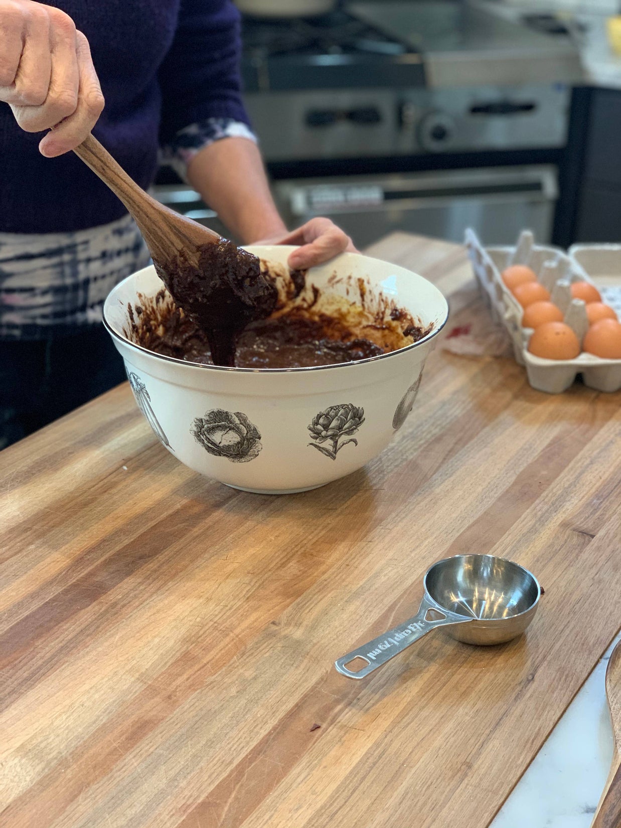 Person mixing chocolate batter in a bowl on a wooden countertop with eggs and measuring cup in the background.