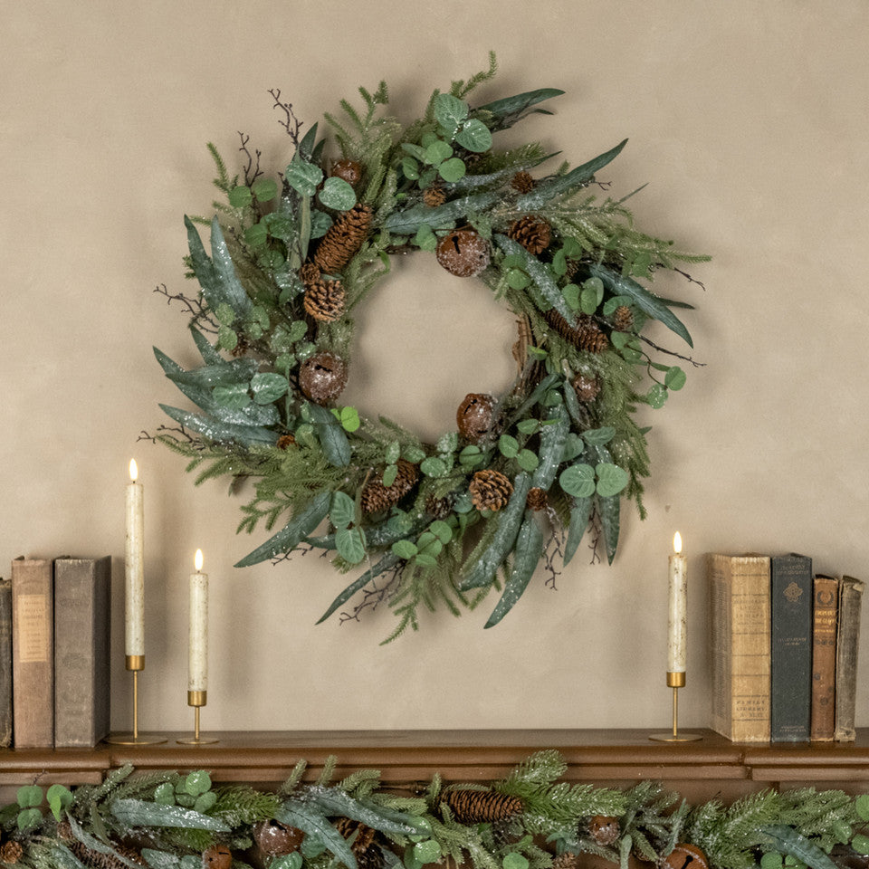 Green wreath with pinecones on a wall above a shelf with books and candles.