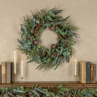 Green wreath with pinecones on a wall above a shelf with books and candles.