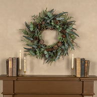 Decorative wreath with greenery and pinecones on a wooden surface with candles and books.