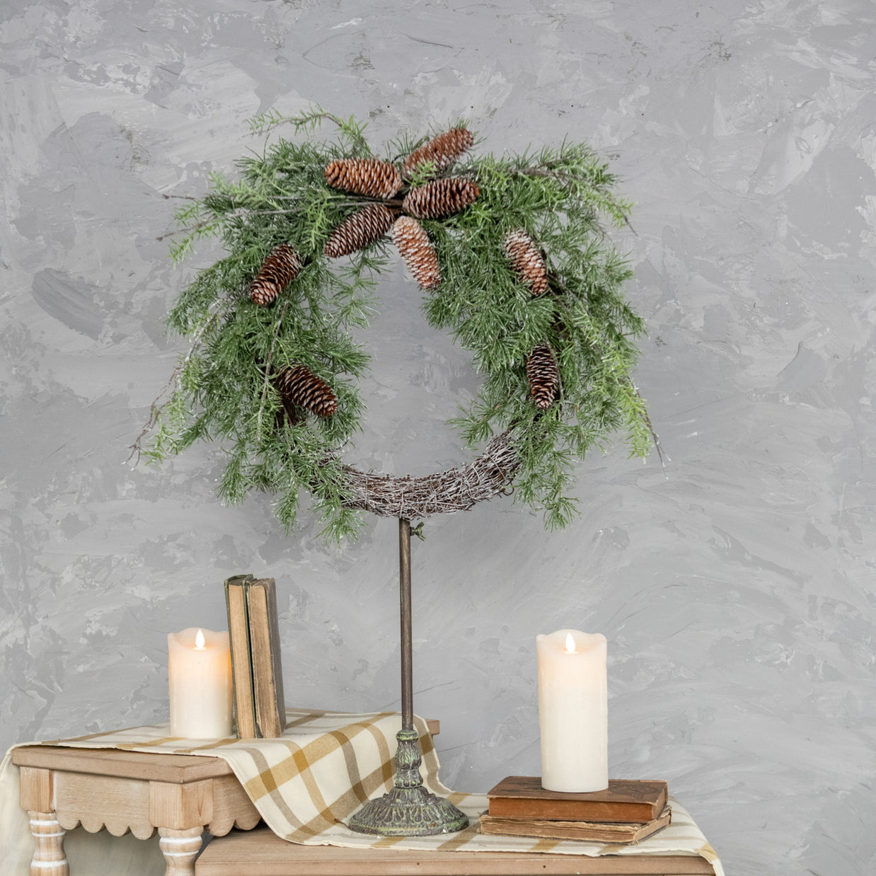 Decorative wreath with pine cones on a stand with candles and books against a gray wall.