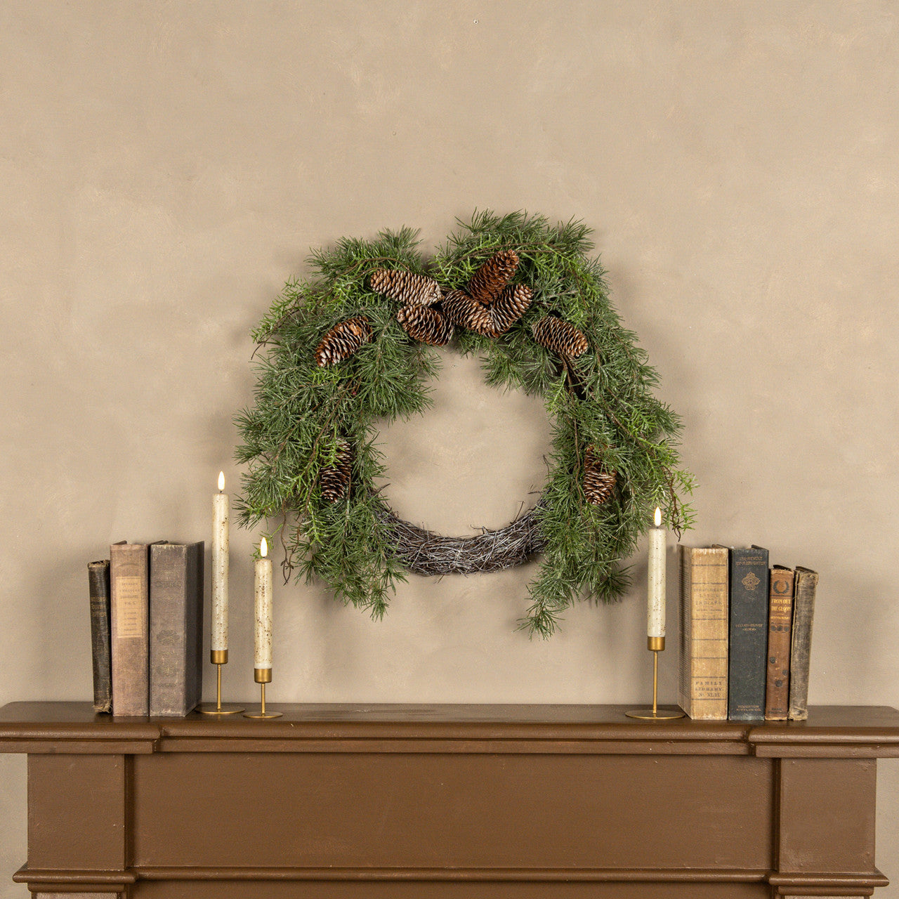 Green wreath with pinecones on a beige wall above a wooden console table with books and candles.