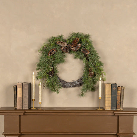 Green wreath with pinecones on a beige wall above a wooden console table with books and candles.