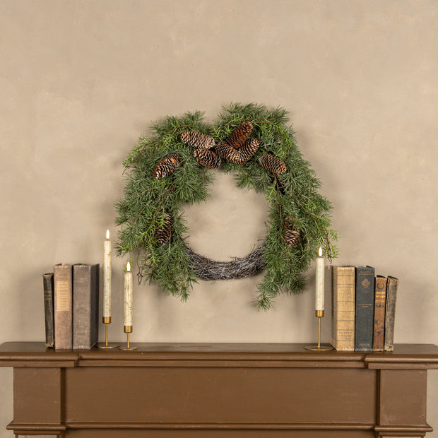 Green wreath with pinecones on a beige wall above a wooden console table with books and candles.