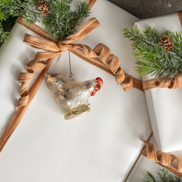 Decorative glass chicken ornament on a wrapped gift with brown ribbon and greenery.