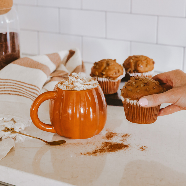 Person holding a muffin next to an orange mug with whipped cream on a kitchen counter.
