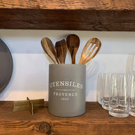 Gray utensil holder with wooden spoons on a wooden shelf with glasses and a decorative item.