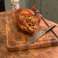 Roasted stuffed chicken on a wooden cutting board with a knife, in a kitchen setting.