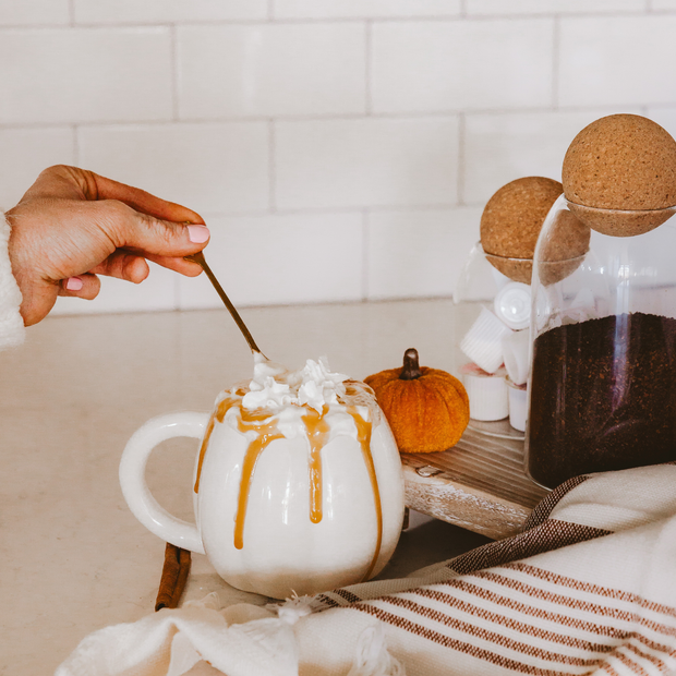 Person stirring a pumpkin spice latte with whipped cream and caramel drizzle in a cozy setting.