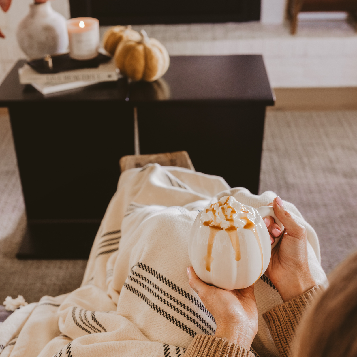 Person holding a small white pumpkin with brown drizzle, sitting on a couch in a cozy living room.