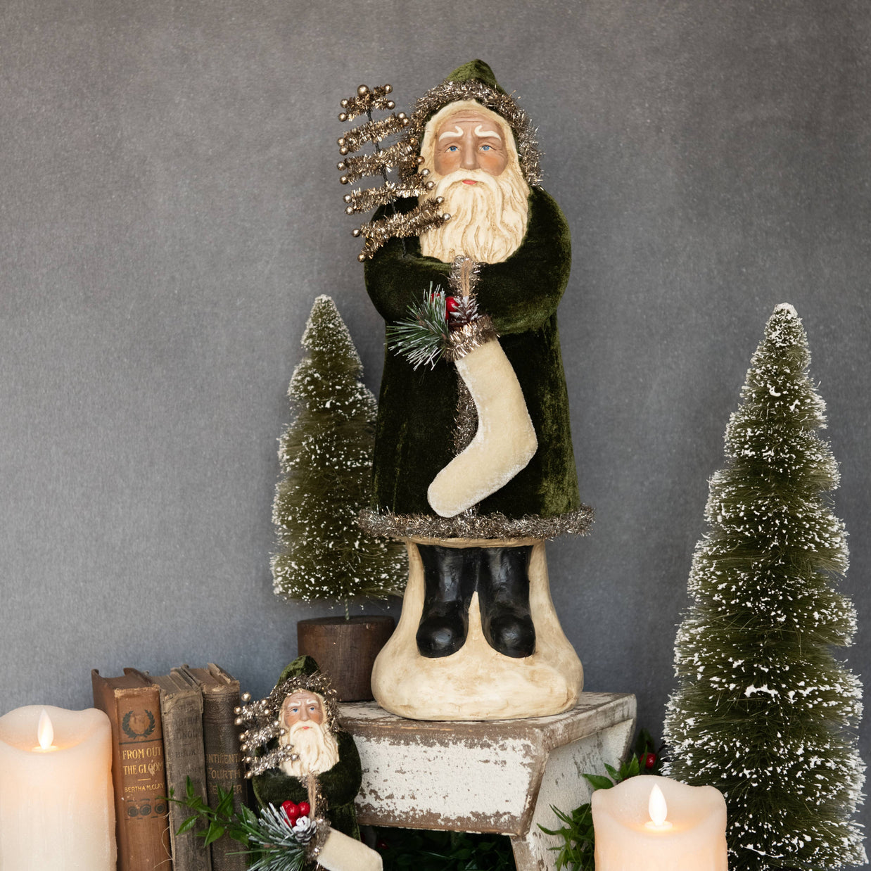 Decorative figure of a bearded man in a green coat with a white beard, holding a branch and a stocking, surrounded by small trees and candles on a gray background.