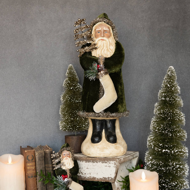 Decorative figure of a bearded man in a green coat with a white beard, holding a branch and a stocking, surrounded by small trees and candles on a gray background.