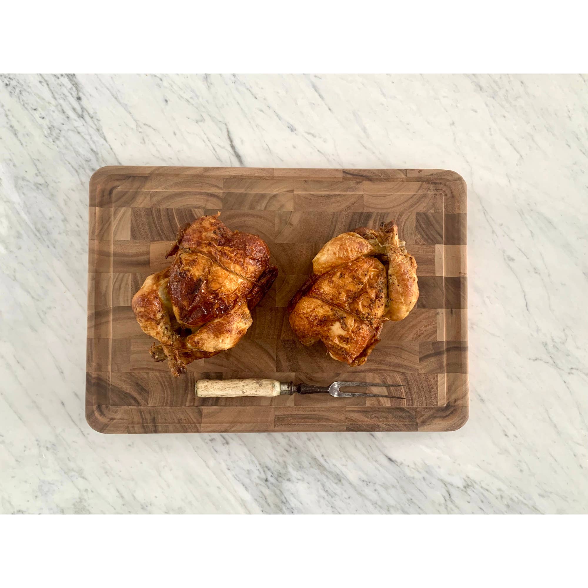 Two roasted chickens on a wooden cutting board with a marble background