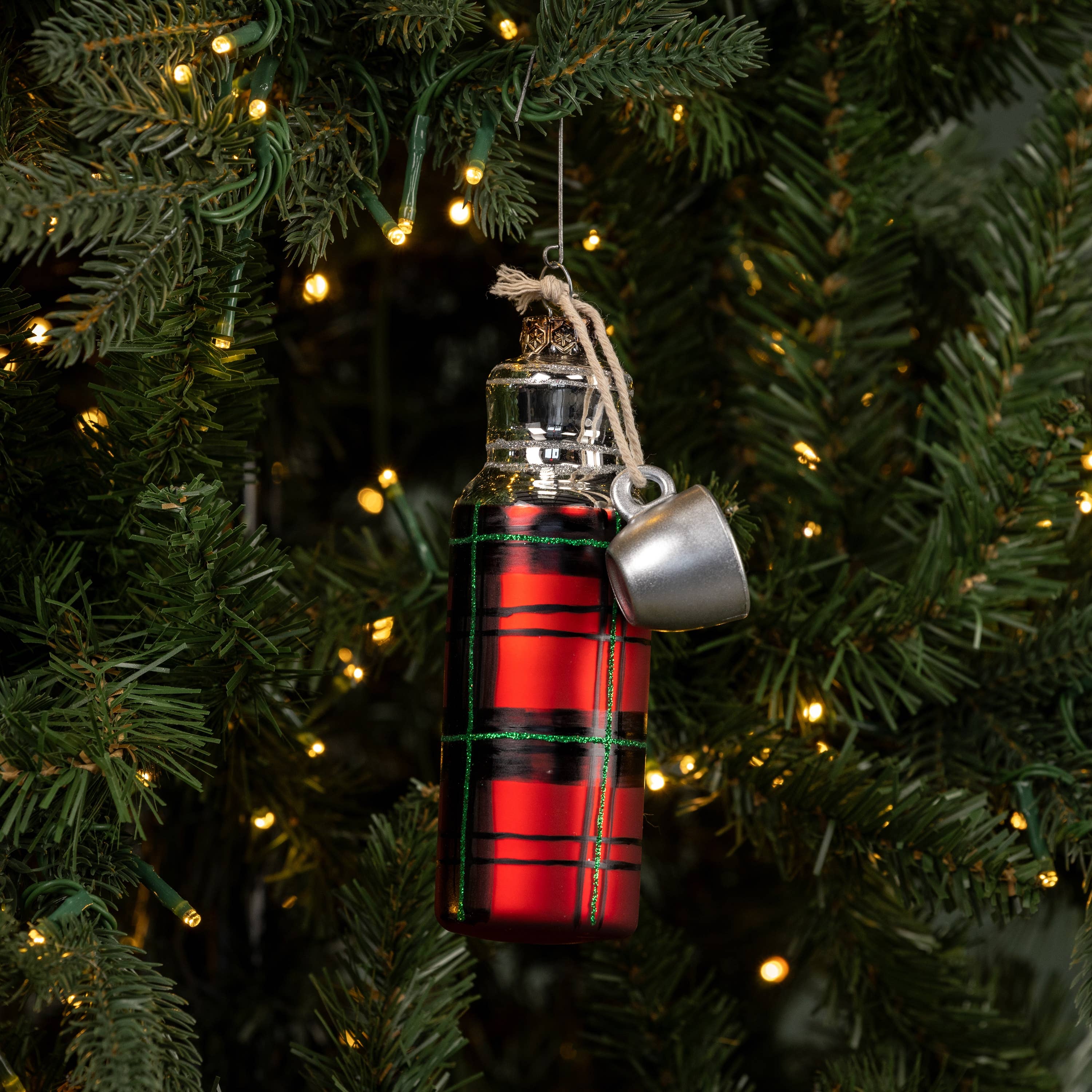 Red plaid bottle ornament with silver top hanging on a Christmas tree.