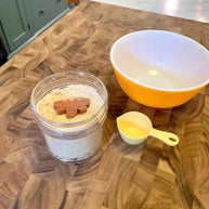 Jar of flour with a bee design, yellow measuring cup, and orange bowl on a wooden surface.