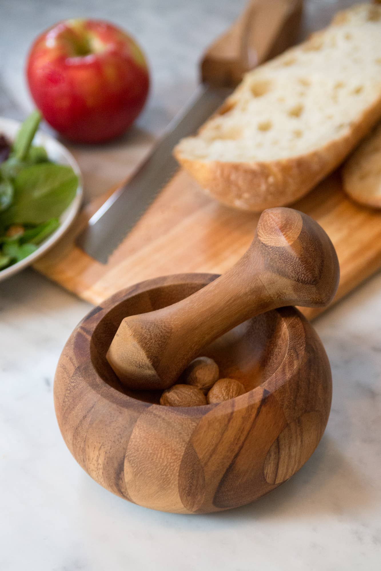 Wooden mortar and pestle with almonds on a marble surface, surrounded by bread, an apple, and salad.