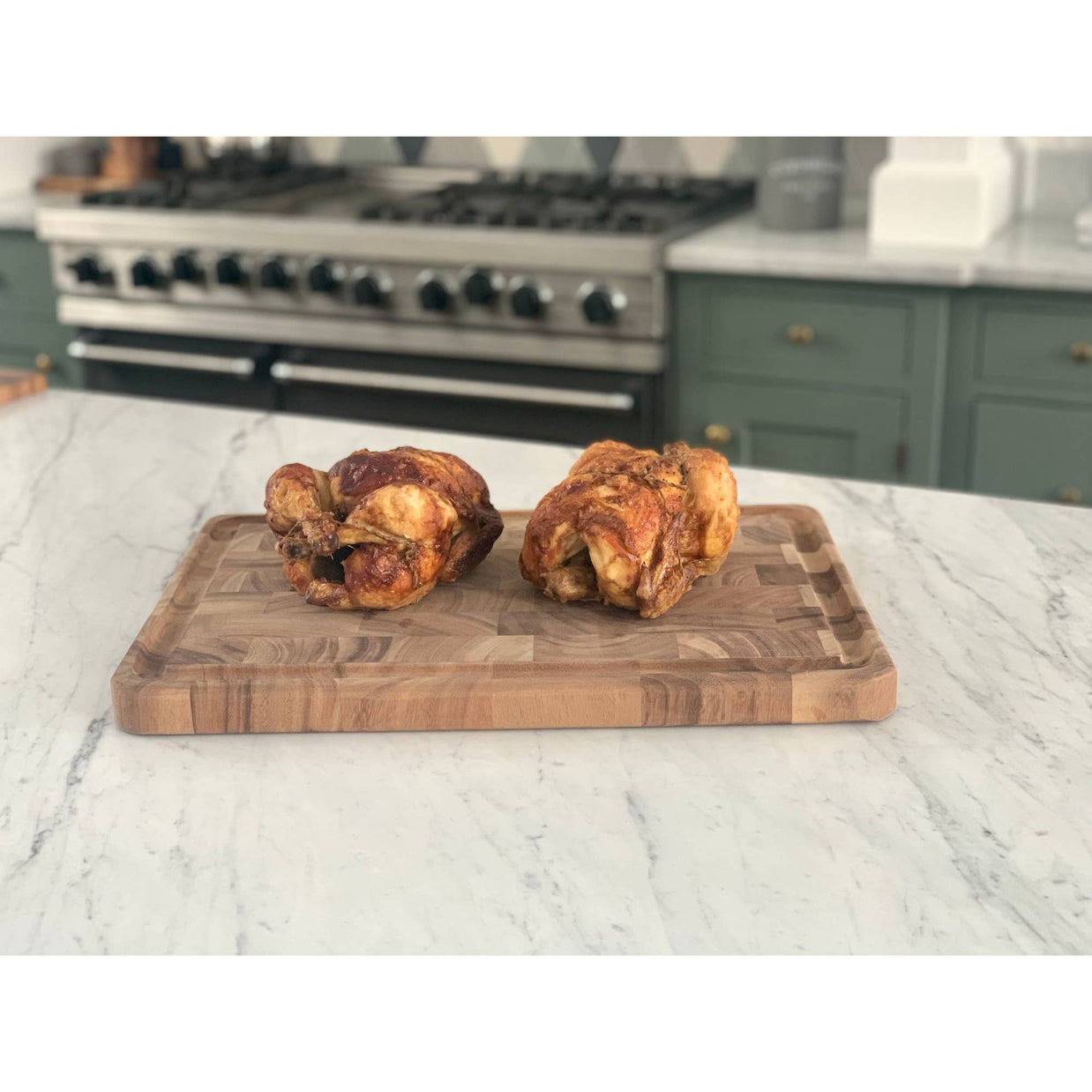 Two roasted chickens on a wooden cutting board in a kitchen setting.