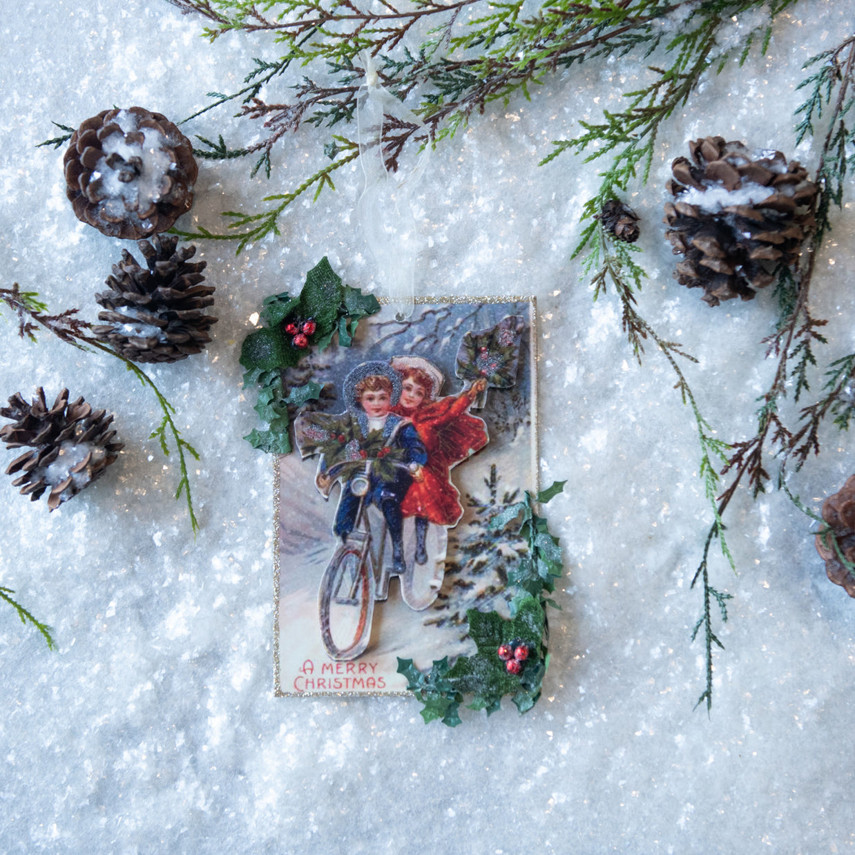 Vintage Christmas card with two children on a bicycle surrounded by pine cones and branches on a snowy background.