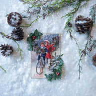 Vintage Christmas card with two children on a bicycle surrounded by pine cones and branches on a snowy background.