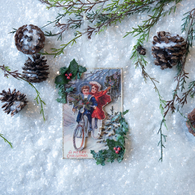 Vintage Christmas card with two children on a bicycle surrounded by pine cones and branches on a snowy background.