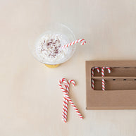 Mug with whipped cream and a candy cane straw on a beige surface, next to a cardboard box.