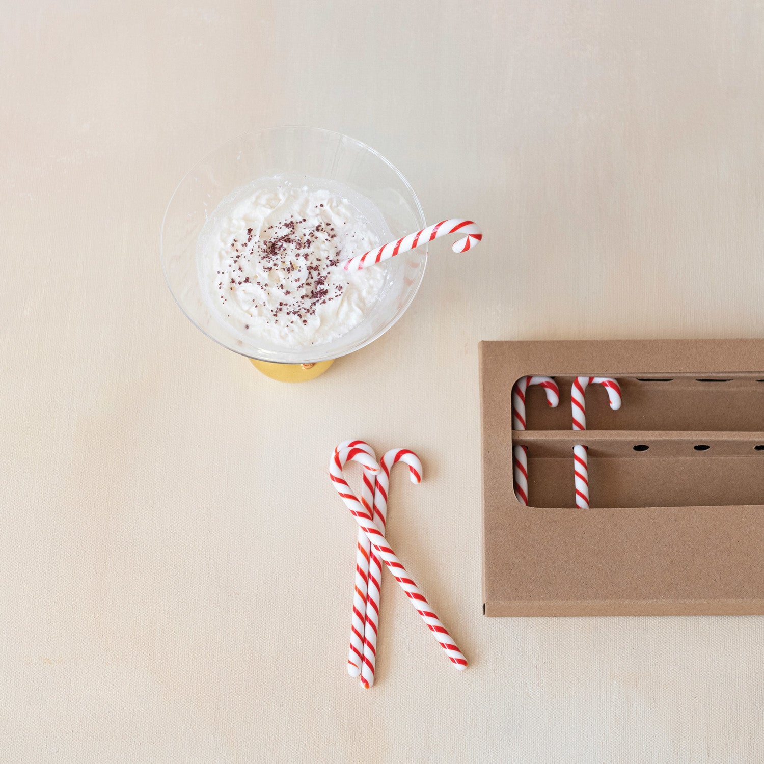 Mug with whipped cream and a candy cane straw on a beige surface, next to a cardboard box.