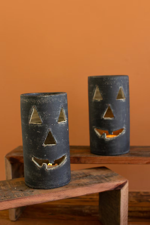 Two black jack-o'-lantern candle holders on a wooden surface against an orange wall.