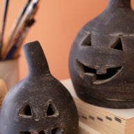 Two black ceramic jack-o'-lanterns on a wooden surface with paintbrushes in the background.