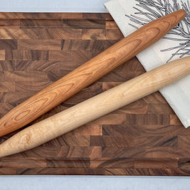 Two wooden rolling pins on a wooden cutting board with a white towel in the background.
