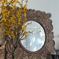 Decorative mirror with intricate carvings next to a potted plant with yellow flowers on a white wall.