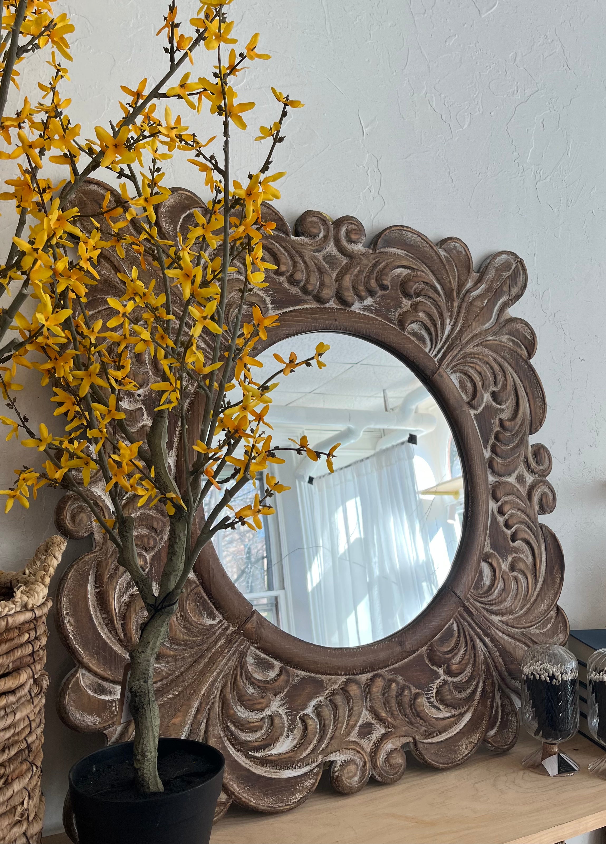Decorative mirror with intricate carvings next to a potted plant with yellow flowers on a white wall.