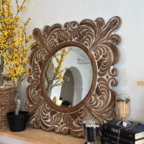 Decorative wall mirror with intricate carvings on a shelf with books and decorative items.