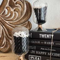 Decorative glass containers with black and white contents on a stack of books against a wooden wall.