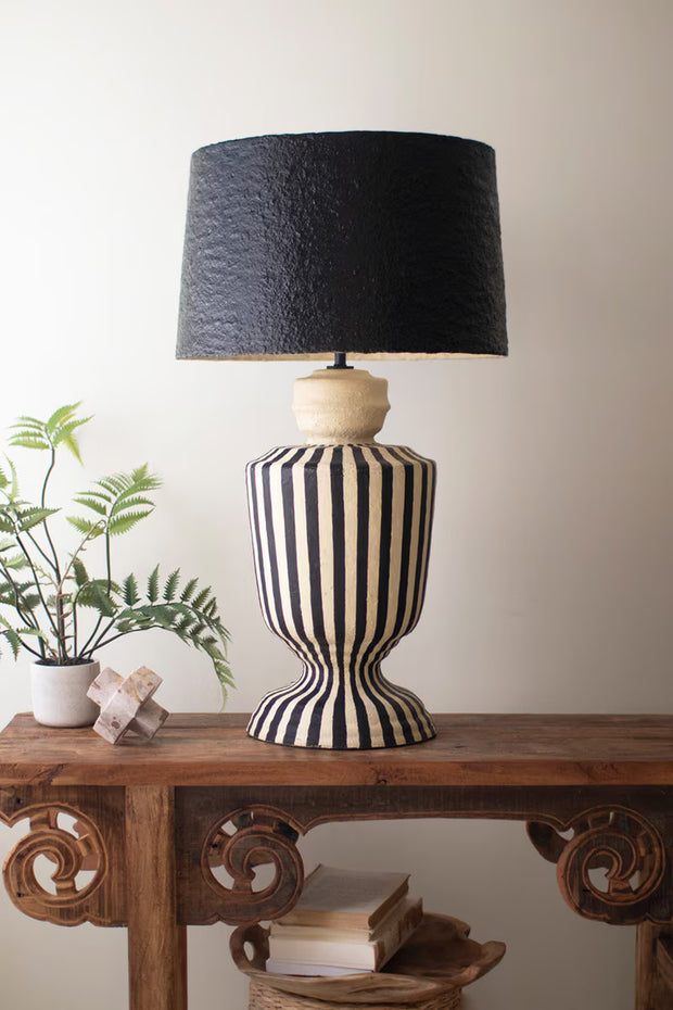 Black and white striped lamp on a wooden console table with a plant and books in the background.