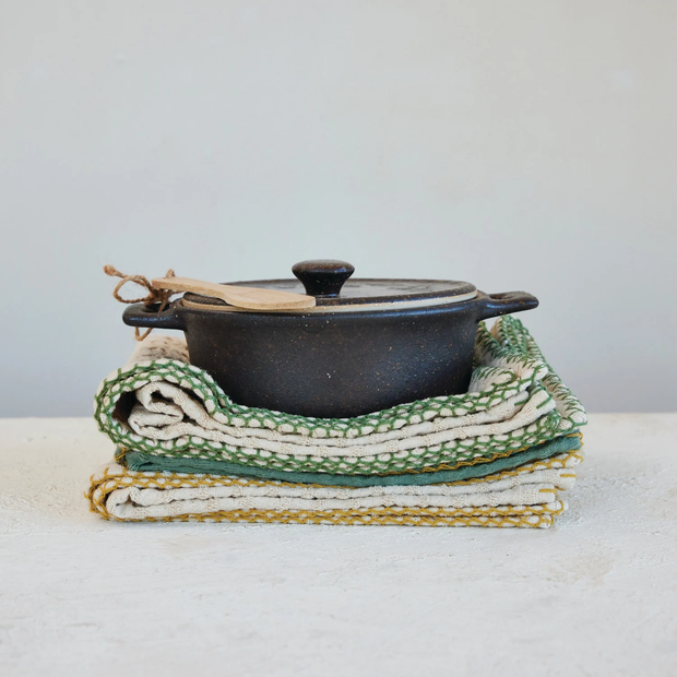 Black pot with a lid on top of folded green and white patterned cloths on a light surface.