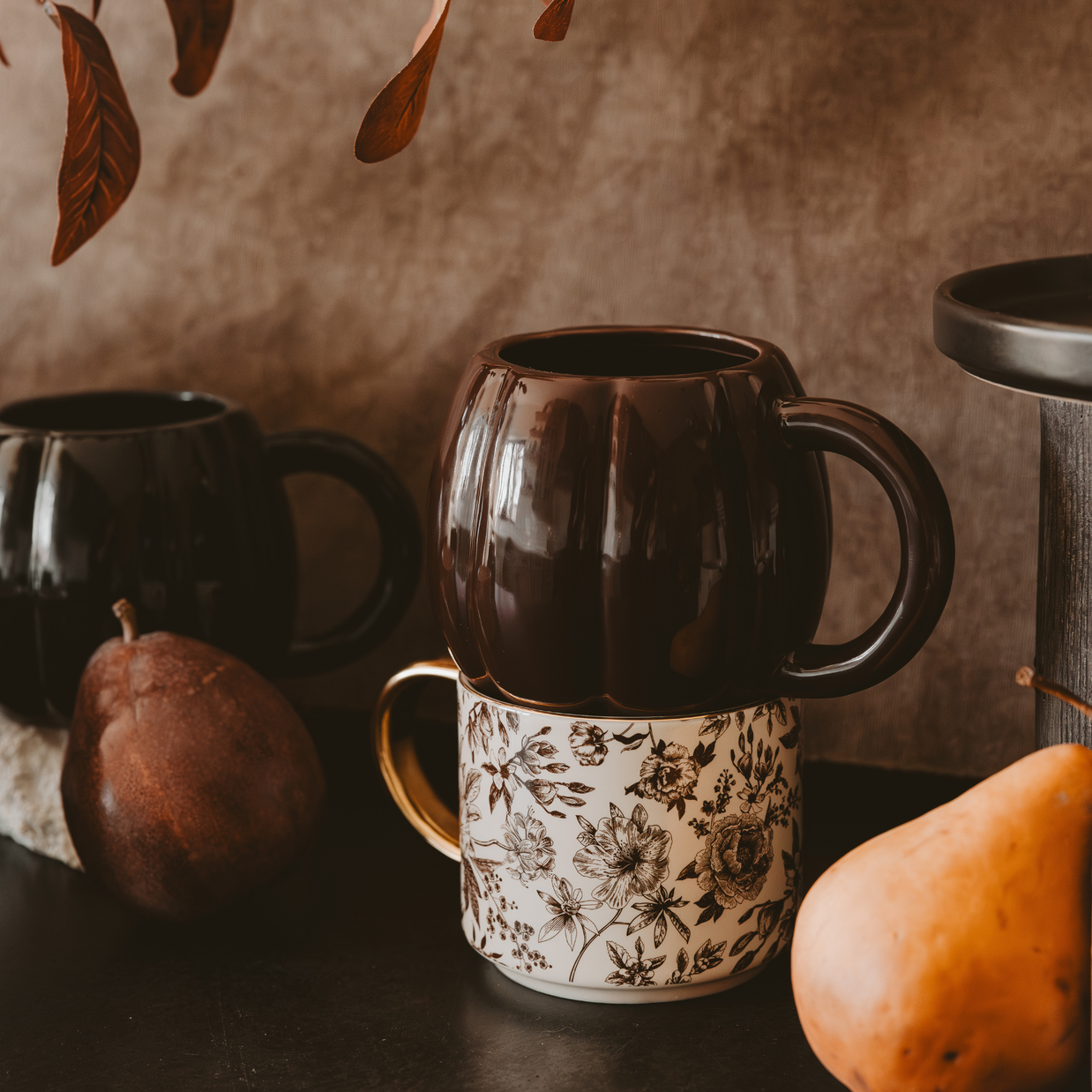 Two black mugs with floral patterns on a dark surface with pumpkins and leaves.