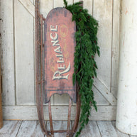 Vintage sled with 'Reliance' branding and greenery against a wooden wall.
