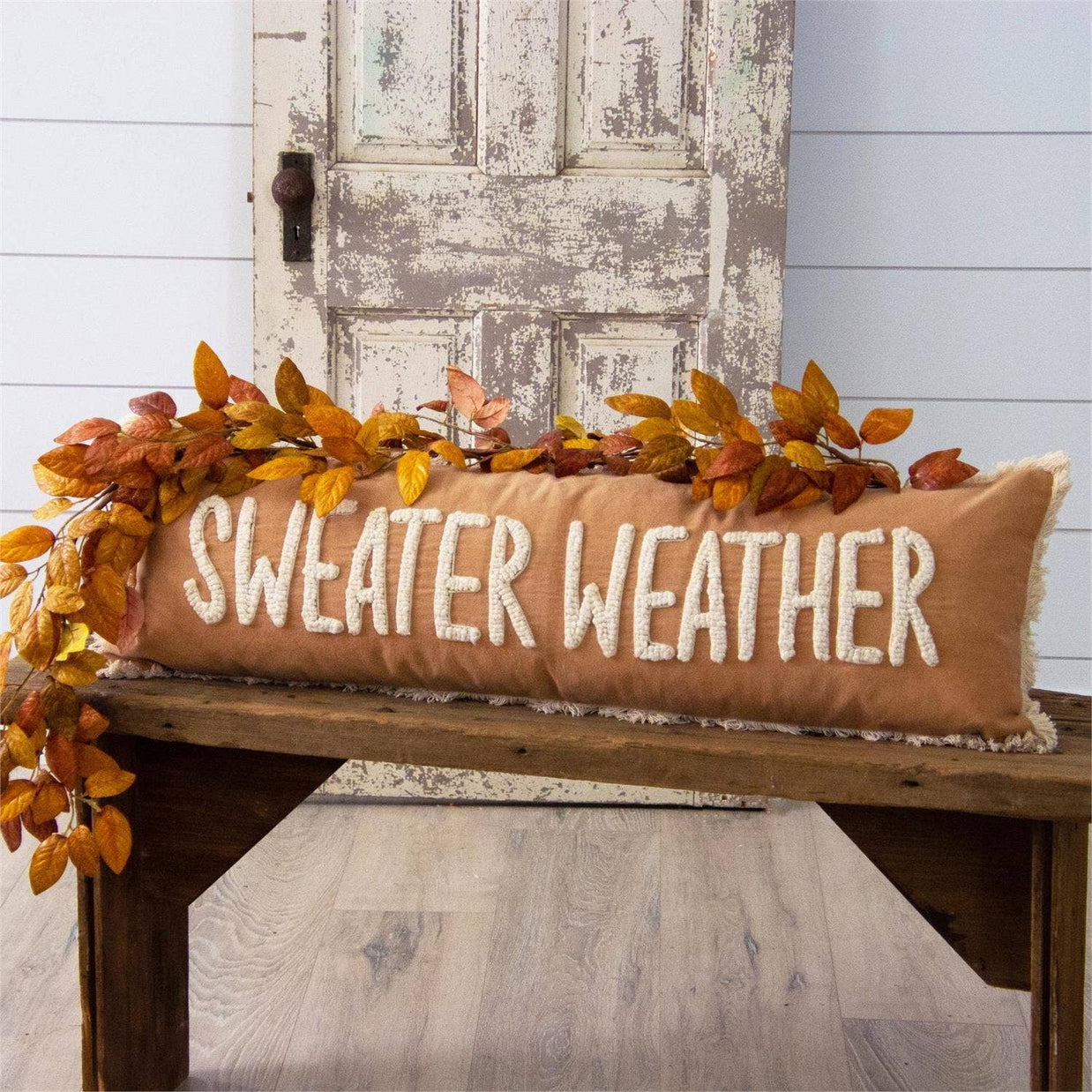Decorative pillow with 'Sweater Weather' text on a wooden bench with autumn leaves.