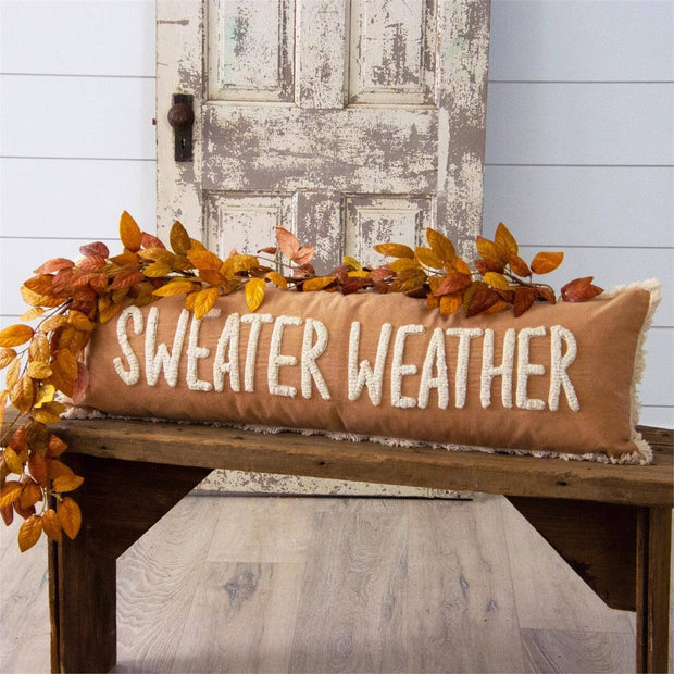 Decorative pillow with 'Sweater Weather' text on a wooden bench with autumn leaves.