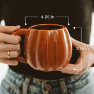 Brown ceramic mug held by hands with measurements indicated, against a blurred background.