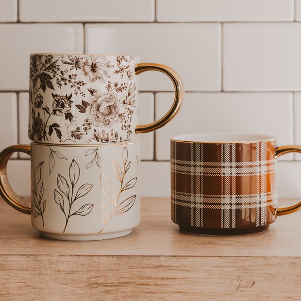 Three ceramic mugs with floral, plaid, and leaf patterns on a wooden surface.