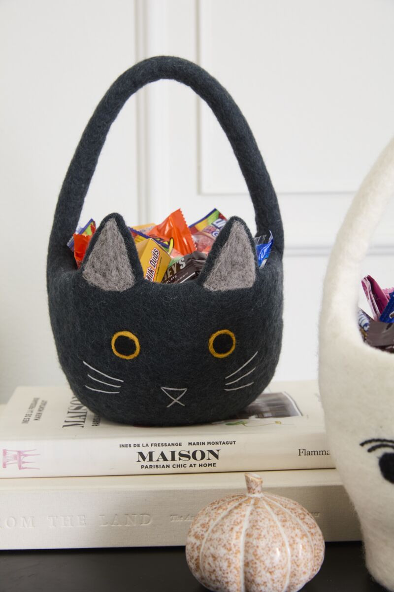 Black cat-shaped felt basket with candy on top of books, next to a white cat-shaped basket.