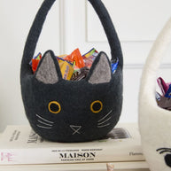 Black cat-shaped felt basket with candy on top of books, next to a white cat-shaped basket.