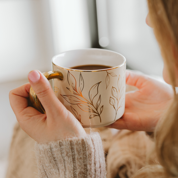 Person holding a white mug with floral design, filled with coffee, against a blurred background.